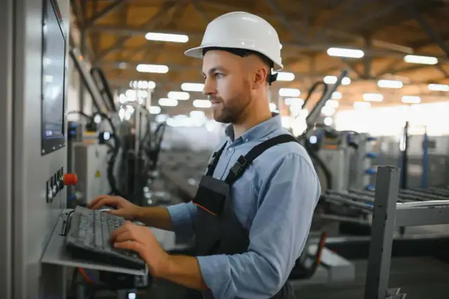 Pracownik w białym kasku obsługuje panel sterowania, wprowadzając dane do systemu. Optymalizacja procesów produkcyjnych dzięki **lean manufacturing**.
