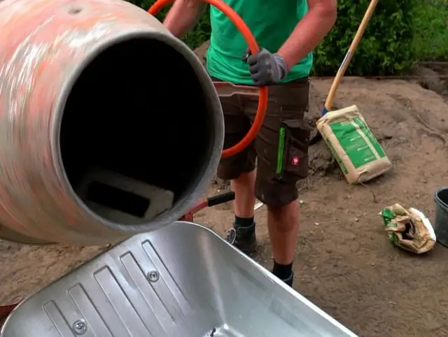 Osoba w rękawiczkach miesza beton w betoniarce, obok stoi taczka i worek cementu.