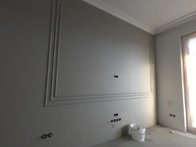 Ściana z ozdobnymi listwami, przygotowana do malowania. Widać wiadro z farbą, co sugeruje, że zaraz nastąpi malowanie agregatem bez smug.