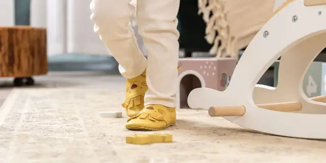Nóżki dziecka w żółtych kapciach z motywem lwa na dywanie obok drewnianej konika na biegunach.