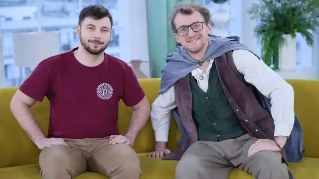 Dwóch mężczyzn, jeden w koszulce, drugi w stroju przypominającym hobbita, siedzi na kanapie.
