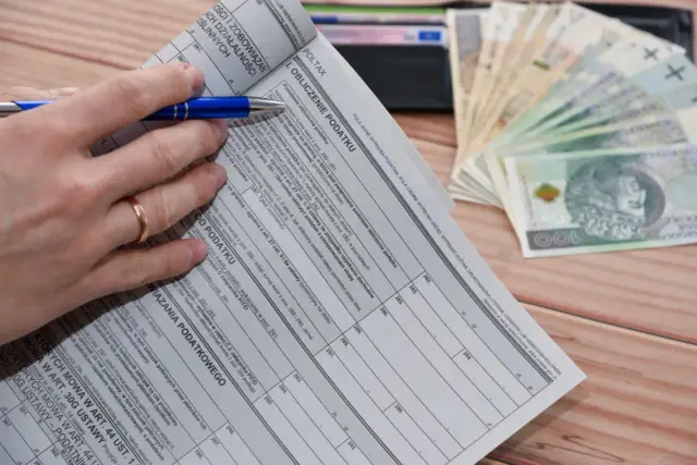 Ręka z długopisem nad deklaracją podatkową, obok stos banknotów. Czas na rozliczenie, kiedy zwrot podatku jest na wyciągnięcie ręki.