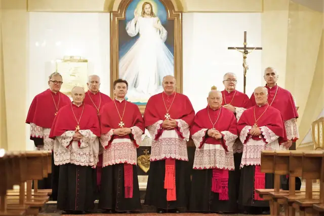 Grupa duchownych w strojach liturgicznych, w tym kilku kanoników, pozuje do zdjęcia przed obrazem Jezusa Miłosiernego.