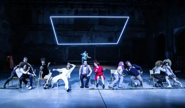 Scena teatralna z grupą ludzi w różnych pozach, jakby w trakcie pantomimy. W tle neonowy prostokąt.