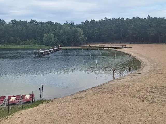 Drewniany pomost nad jeziorem Kamionki, piaszczysta plaża, łódki i dwie osoby kąpiące się w wodzie.