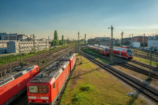 Czerwone lokomotywy i wagony stoją na torach. W tle budynki i drzewa. To miejsce, gdzie przygotowuje się kolej dużych prędkości do podróży.