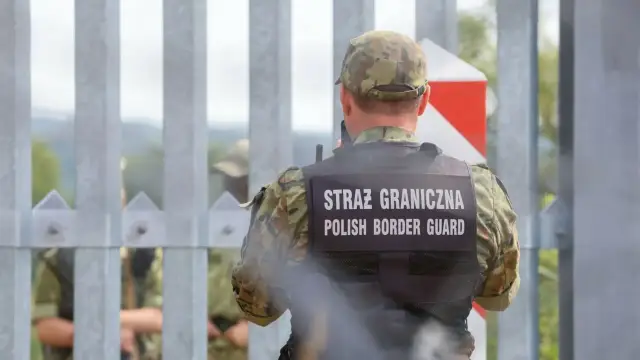 Strażnik graniczny w kamizelce z napisem "STRAŻ GRANICZNA POLISH BORDER GUARD" stoi przy płocie. W tle widać innych funkcjonariuszy i znak graniczny. To obraz polskiej ochrony granic z Rosją.