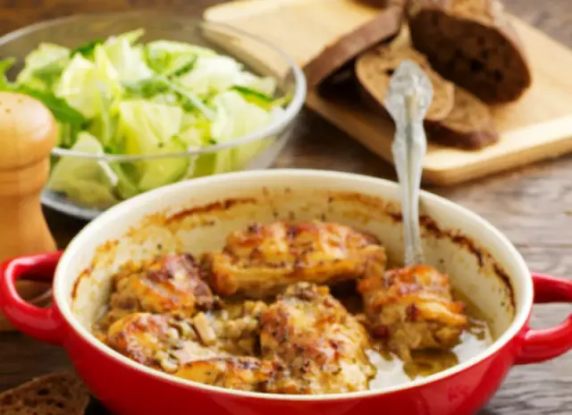Pieczony królik w czerwonym naczyniu, z dodatkiem marynaty do królika pieczonego w piekarniku. Obok sałatka i chleb.