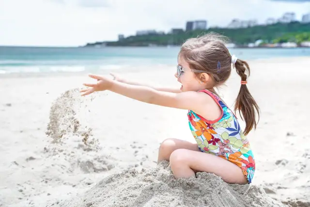Dziewczynka w kolorowym stroju kąpielowym z okularami na nosie rzuca piasek, ciesząc się zabawami na plaży.
