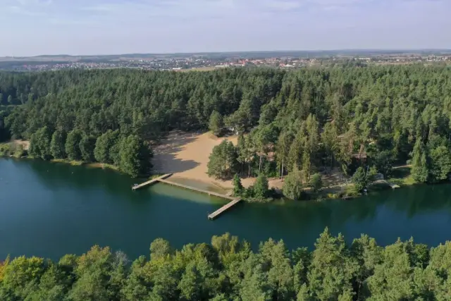 Malownicza borówno plaża otoczona lasem, z drewnianymi pomostami wchodzącymi do ciemnej wody. W oddali widać zabudowania.