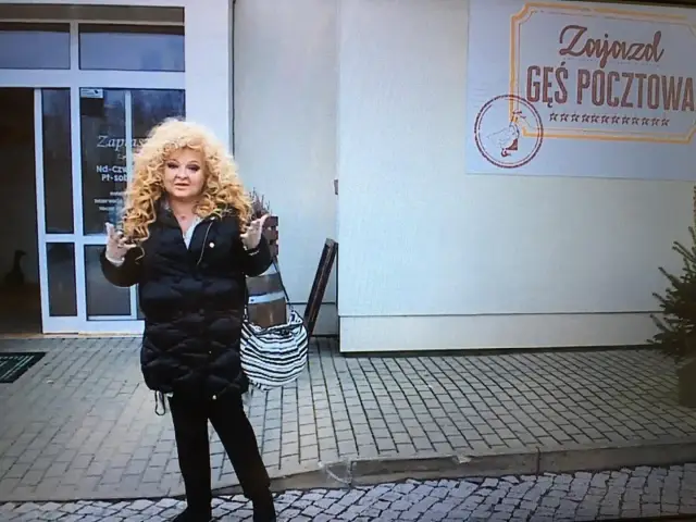 Kobieta z burzą blond włosów stoi przed Zajazdem Gęś Pocztowa.
