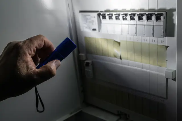 Ręka trzymająca niebieską latarkę oświetla rozdzielnicę elektryczną, pomagając sprawdzić, gdzie jest awaria prądu.