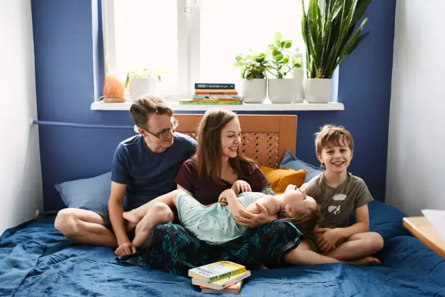 Rodzicielstwo bliskości w praktyce: rodzina śmieje się i przytula na łóżku. Mama tuli córeczkę, tata i starszy syn patrzą z uśmiechem.
