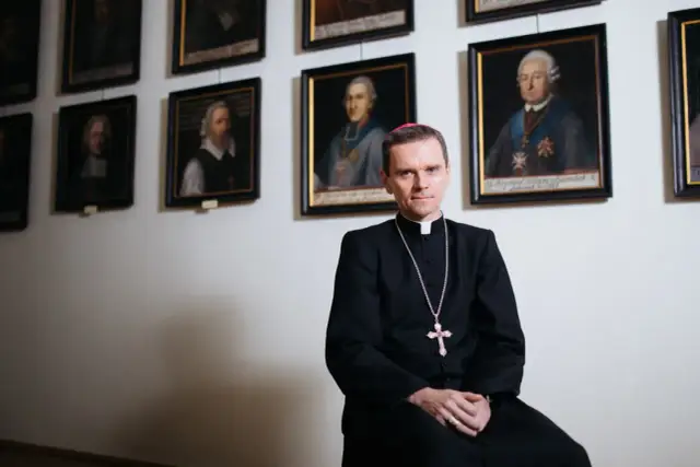 Mężczyzna w stroju duchownym siedzi na tle ścian z portretami.