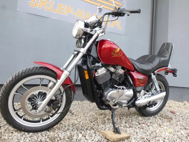 Czerwony motocykl Honda Shadow 500 z chromowanymi elementami, zaparkowany na żwirze przed szarym budynkiem.