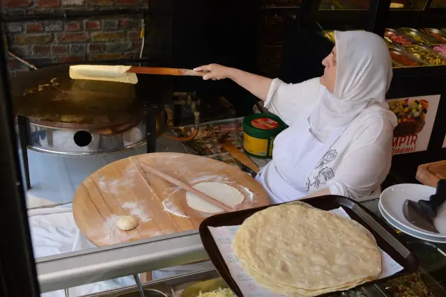 Kobieta w chuście przygotowuje tureckie jedzenie, smażąc cienkie placki na okrągłej płycie. Obok leżą gotowe placki i ciasto.
