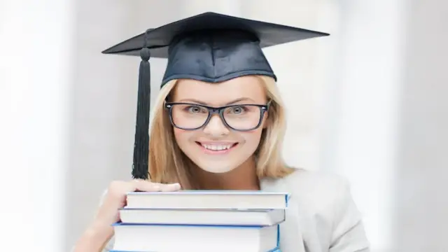 Uśmiechnięta studentka w czapce absolwenta i okularach trzyma stos książek, zastanawiając się, ile trwają studia podyplomowe.
