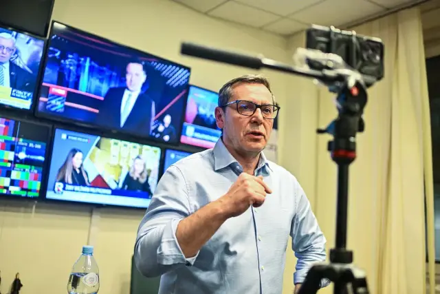 Prezes TVP w okularach, gestykuluje przed kamerą. W tle monitory z programami informacyjnymi.