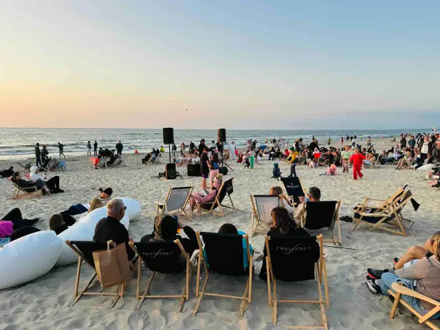 Wieczorne **krynica morska imprezy na plaży**. Ludzie siedzą na leżakach, słuchając muzyki przy zachodzącym słońcu.