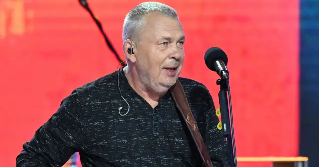 Adam Nowak śpiewa do mikrofonu, ubrany w czarną koszulę, z gitarą na ramieniu.