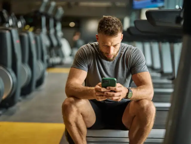 Mężczyzna w siłowni sprawdza telefon, szukając informacji o tym, co spala najwięcej kalorii. Tabela z ćwiczeniami jest w jego myślach.