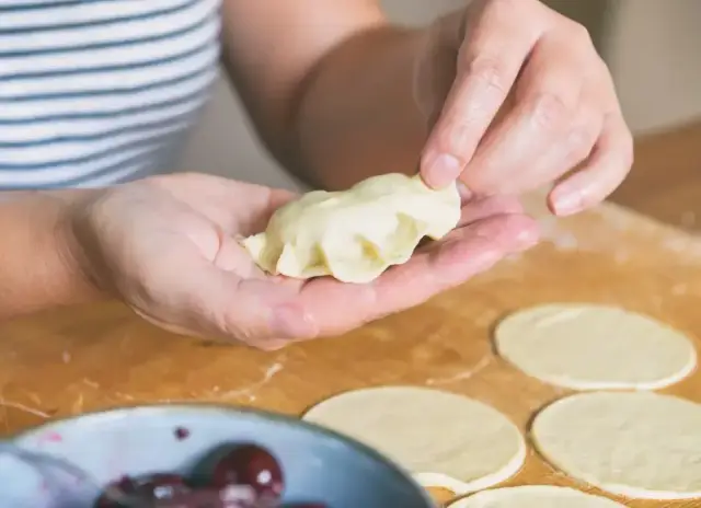 Dłonie formują pieroga z ciasta na pierogi bez jajka. Na blacie leżą okrągłe placki z ciasta i miseczka z wiśniami.