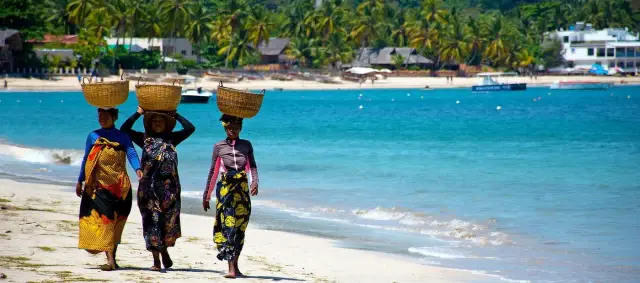 Trzy kobiety niosą kosze na głowach na plaży. Tropikalne palmy i budynki w tle. Idealne wakacje na Madagaskarze.