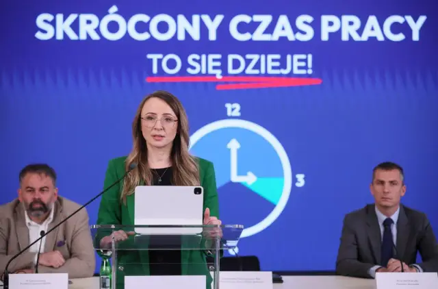 Kobieta przemawia na tle hasła "SKRÓCONY CZAS PRACY TO SIĘ DZIEJE!". Obok niej zegar symbolizujący skrócenie czasu pracy.