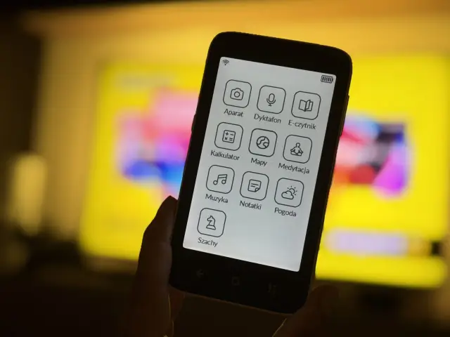 Smartfon z ikonami aplikacji: aparat, dyktafon, e-czytnik, kalkulator, mapy, medytacja, muzyka, notatki, pogoda, szachy. Tło to rozmyty ekran telewizora.