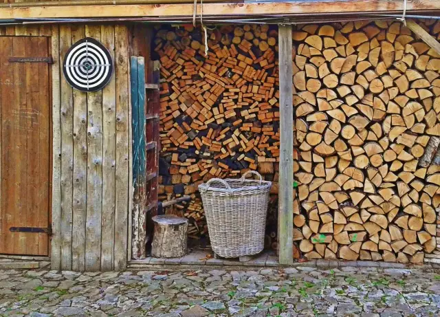 Drewno kominkowe ułożone w szopie, obok kosz i tarcza do rzutek. Widać różne sposoby, jak układać drewno kominkowe, by było suche i gotowe do palenia.