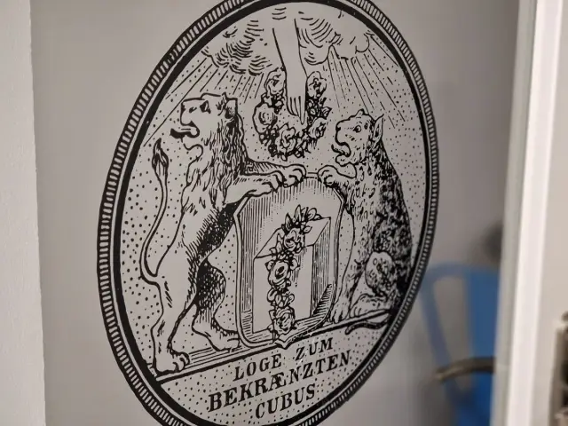 Herb z lwem i panterą, symbolizujący masonerię. Pod spodem napis "LOGE ZUM BEKRÄNZTEN CUBUS".