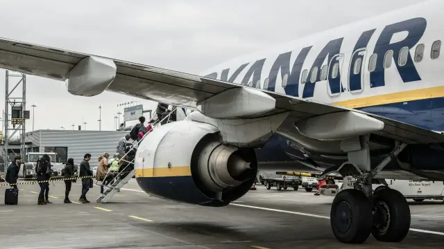 Pasażerowie z bagażem wchodzą po schodkach do samolotu Ryanair. Słońce przebija się przez chmury, zapowiadając podróż.
