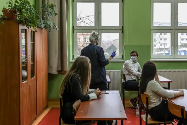 Nauczyciel dyplomowany zarobki - lekcja w klasie, uczniowie w maskach siedzą przy ławkach, nauczyciel trzyma kartkę.