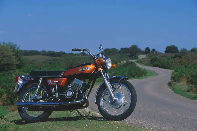 Pomarańczowy motocykl Yamaha RD 350 na tle krętej drogi i zielonych wzgórz. Klasyk gotowy na podróż.