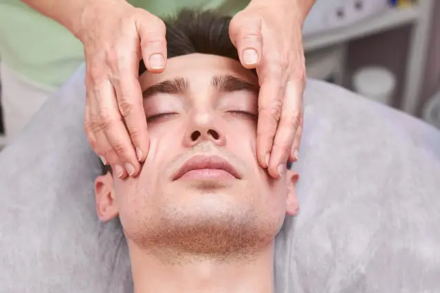 Mężczyzna z zamkniętymi oczami doświadcza relaksującego masażu twarzy podczas kursu Kobido. Dłonie terapeuty delikatnie głaszczą jego skórę.