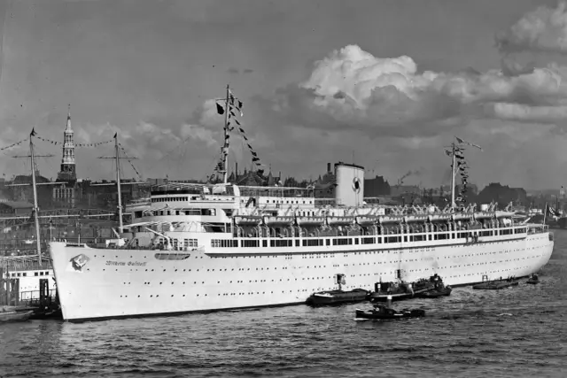 Ogromny statek pasażerski Wilhelm Gustloff cumuje w porcie, otoczony mniejszymi łodziami. W tle widoczne miasto z wieżami kościołów.