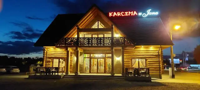 Drewniana karczma u Jana o zmierzchu, oświetlona ciepłym światłem. Neonowy szyld z nazwą lśni nad wejściem.