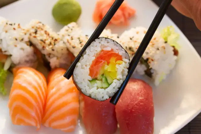 Chopsticks trzymają rolkę sushi z łososiem i warzywami. Na talerzu widać nigiri z łososiem i tuńczykiem, a także inne rolki. Idealna zaprawa do sushi czeka na skosztowanie.