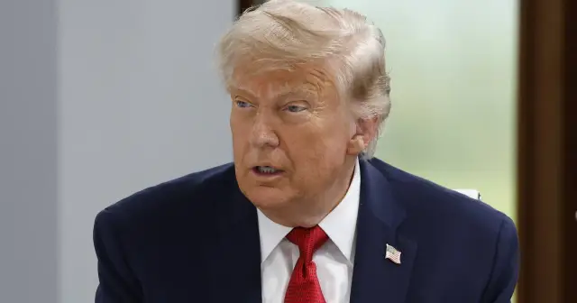 Donald Trump w garniturze i czerwonym krawacie, z amerykańską flagą na klapie, szuka porozumienia.