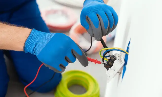 Elektryk w niebieskich rękawicach sprawdza przewody przy gniazdku. Pokazuje, jak podłączyć gniazdko bez uziemienia, używając miernika.
