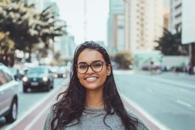 Uśmiechnięta młoda kobieta w okularach na tle miejskiej ulicy. Jej opis postaci po angielsku to "Smiling young woman with glasses on a city street.