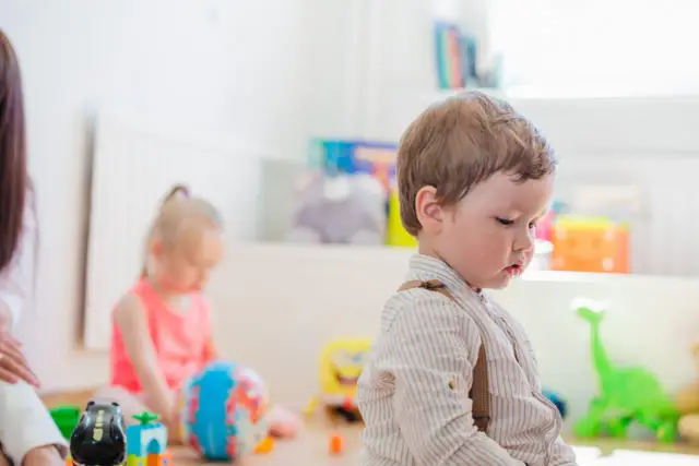 Chłopiec w żłobku, zastanawia się, czy można zapisać dziecko do żłobka w trakcie roku. W tle inne dzieci i zabawki.