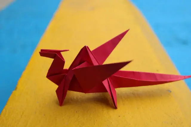 Czerwony smok z papieru, piękne origami z papieru, siedzi na żółtej powierzchni obok niebieskiego tła.
