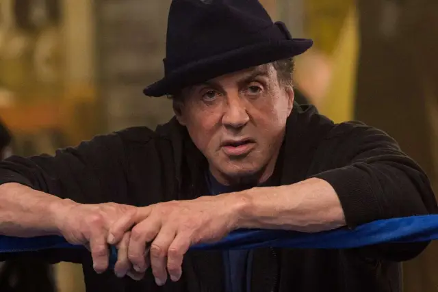 Sylvester Stallone jako Rocky Balboa w czarnym kapeluszu i bluzie, oparty o ring bokserski.