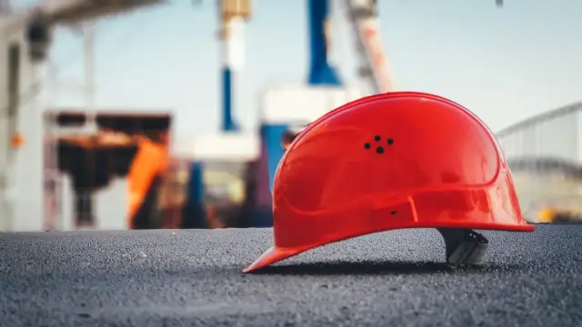 Czerwony kask na budowie symbolizuje ochronę przed ryzykiem zawodowym. W tle widać konstrukcje i sprzęt budowlany.