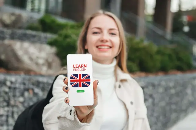 Uśmiechnięta dziewczyna trzyma telefon z napisem "LEARN ENGLISH" i flagą Wielkiej Brytanii. To darmowa aplikacja do nauki angielskiego.