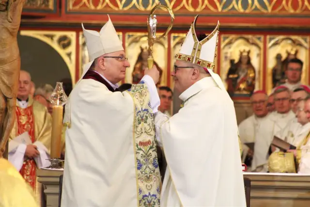 Dwóch biskupów w białych szatach i mitrach podczas ingresu do katedry. Jeden przekazuje drugiemu pastorał.