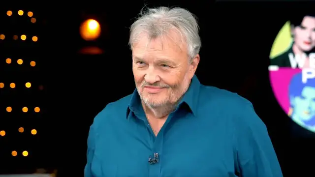 Krzysztof Cugowski, w niebieskiej koszuli, z siwą brodą i włosami, uśmiecha się. W tle migoczą światła, przypominając o jego wspaniałych utworach.