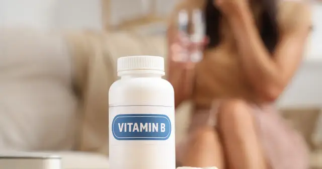Biała butelka z etykietą "VITAMIN B" stoi na stole. W tle kobieta pije wodę. Tiamina, składnik witaminy B, wspiera metabolizm.