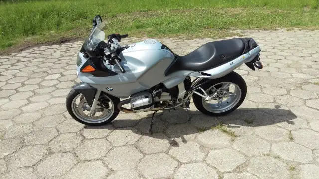 Srebrny motocykl BMW R1100S na betonowym placu, gotowy do jazdy po zielonych terenach.